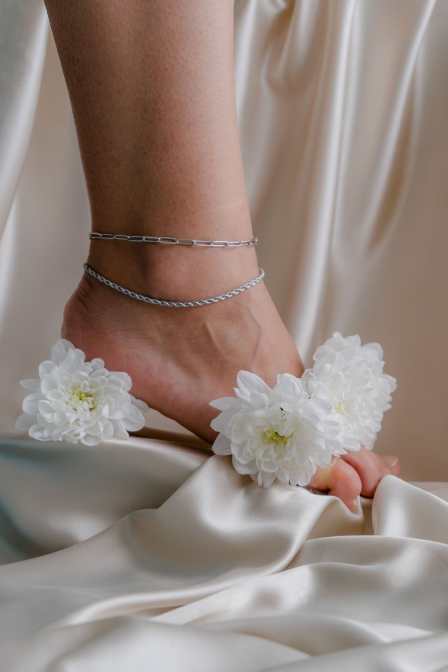 Delicate anklet chains on an ankle resting on pale fabric with white flowers